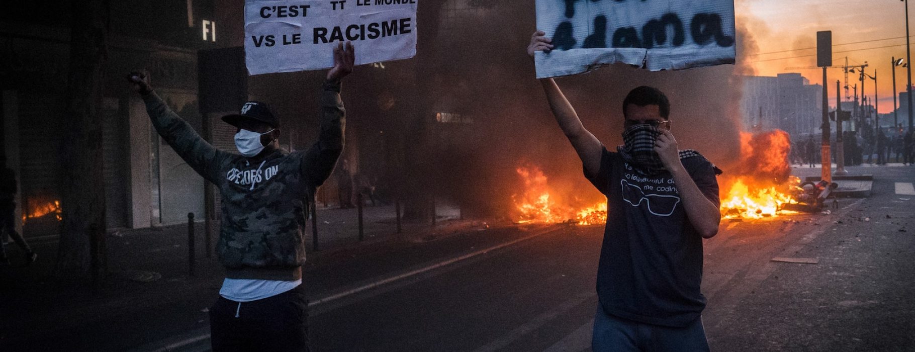 Two people in masks hold up signs in front of a fire.